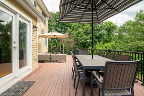 Relax on the outdoor deck, surrounded by lush greenery, with a dining table, wicker sofa, and umbrella for shade.