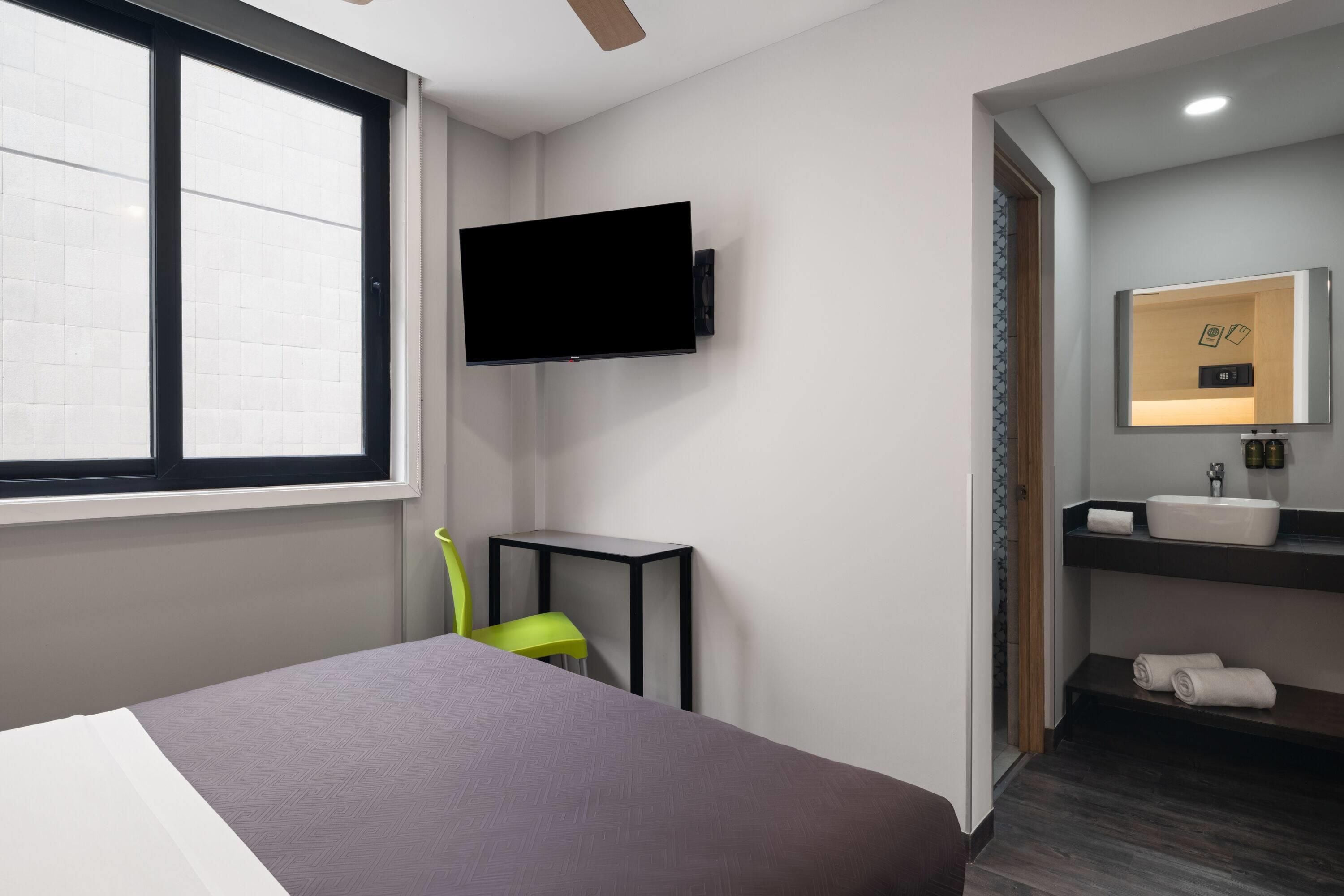 Ein modernes Hotelzimmer mit einem Bett, einem an der Wand befestigten Fernseher, einem kleinen Tisch, einem Sessel und einem Badezimmer mit Waschbecken und Spiegel.