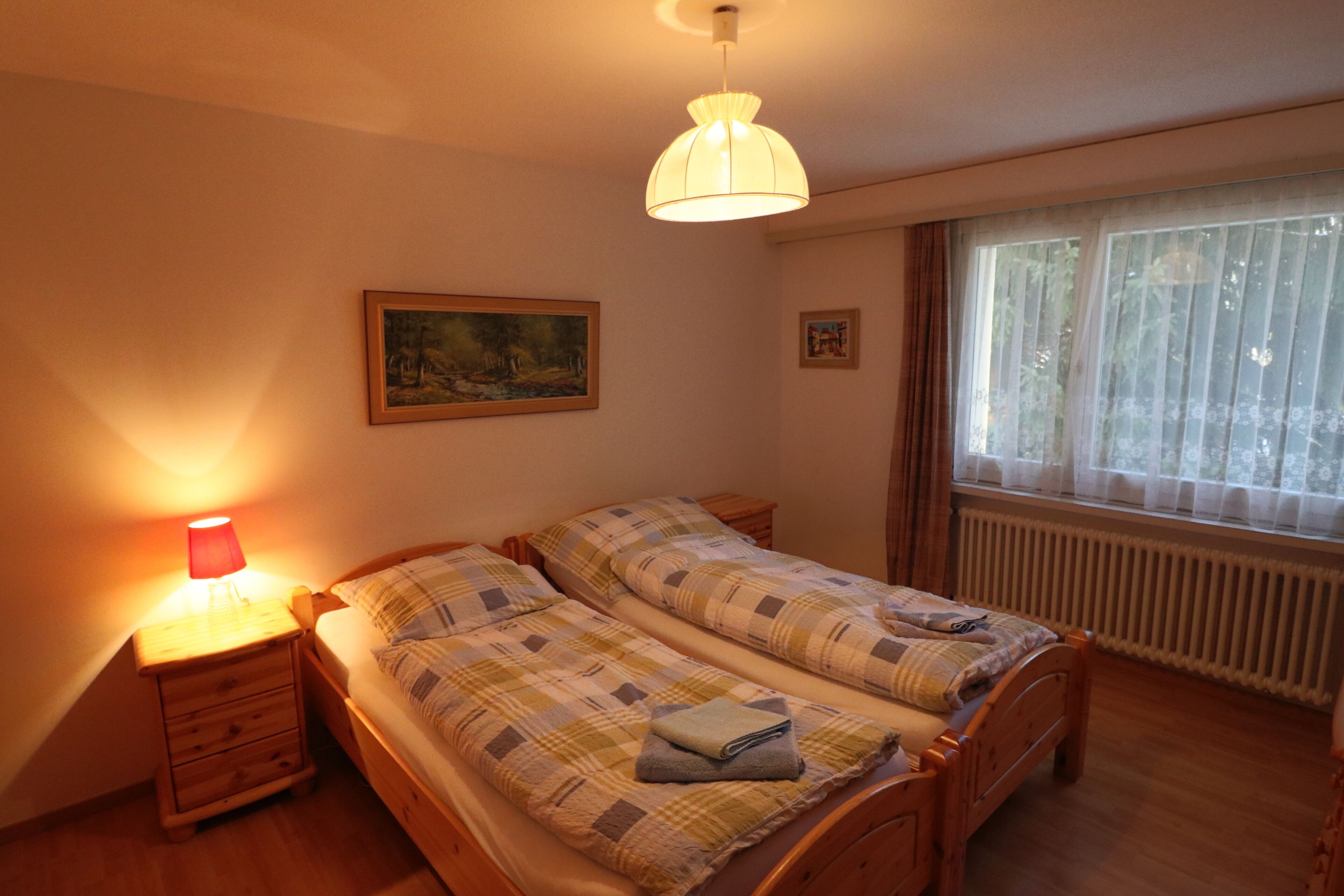 A bedroom with two beds, a nightstand, a lamp, a painting, and a window with curtains.