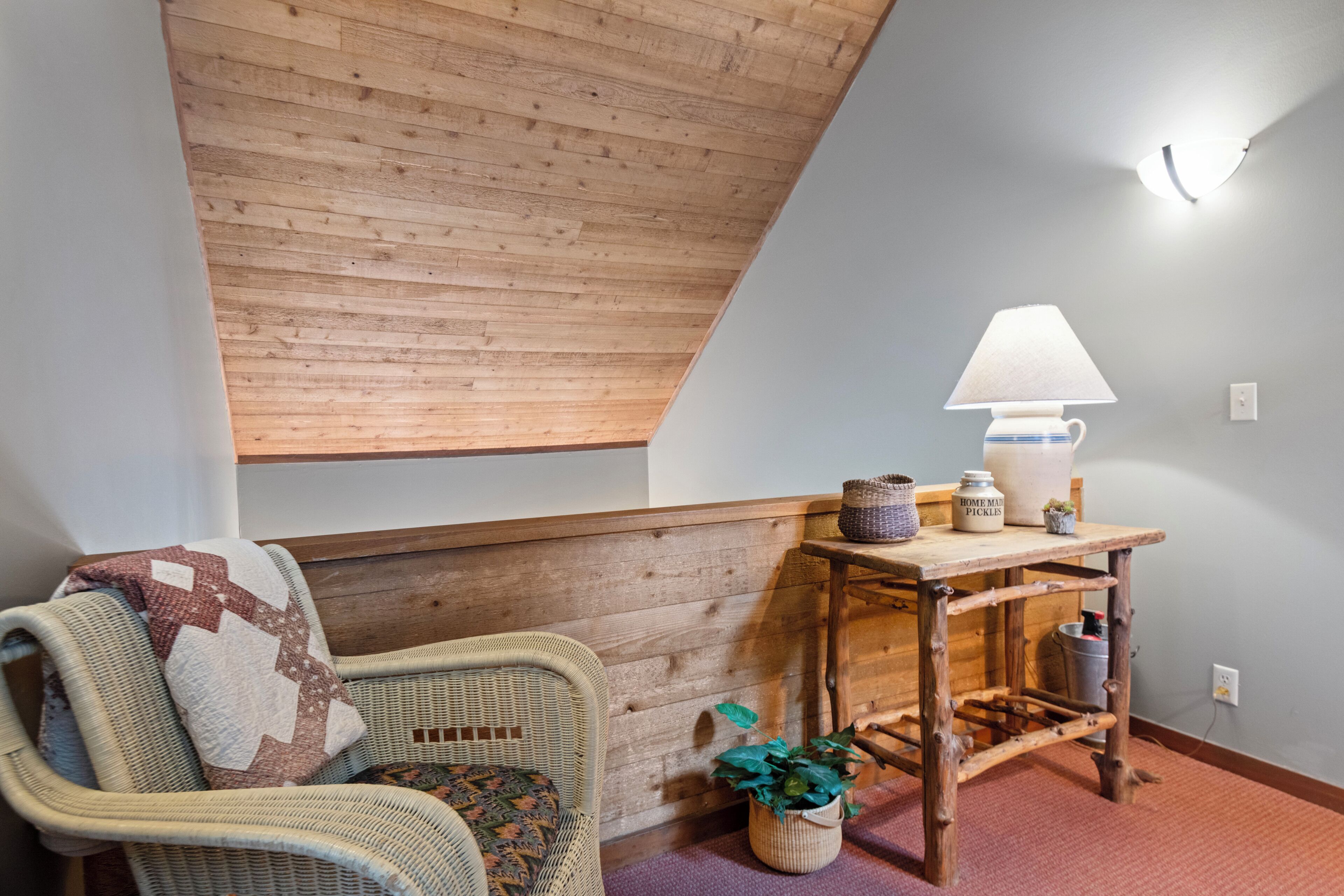 Een knus vertrek met een schuine kap, een rieten stoel met een geborduurd plaid, een houten bijzettafeltje met een lamp, en een kleine potplant.