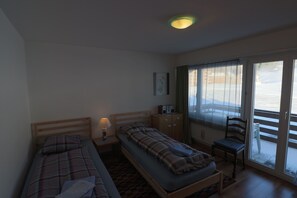 A room with two single beds, a chair, a small table, a radio, and a window with curtains. - Apartments Museumstrasse (Davos)
