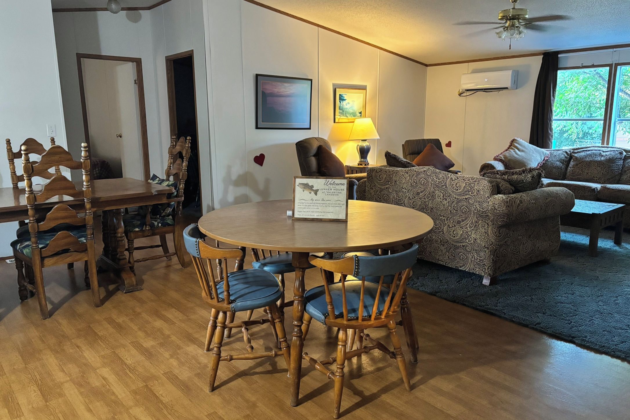 A cozy living room with a dining table, chairs, a sofa, and a coffee table. There is a sign on the wall and a ceiling fan.
