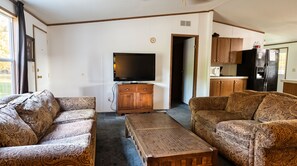 A living room with patterned sofas, a television, a wooden cabinet, and a kitchen area with a refrigerator.