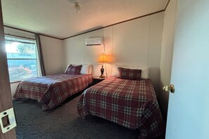 A room with two beds, a nightstand with a lamp, a window with a view, and an air conditioning unit.