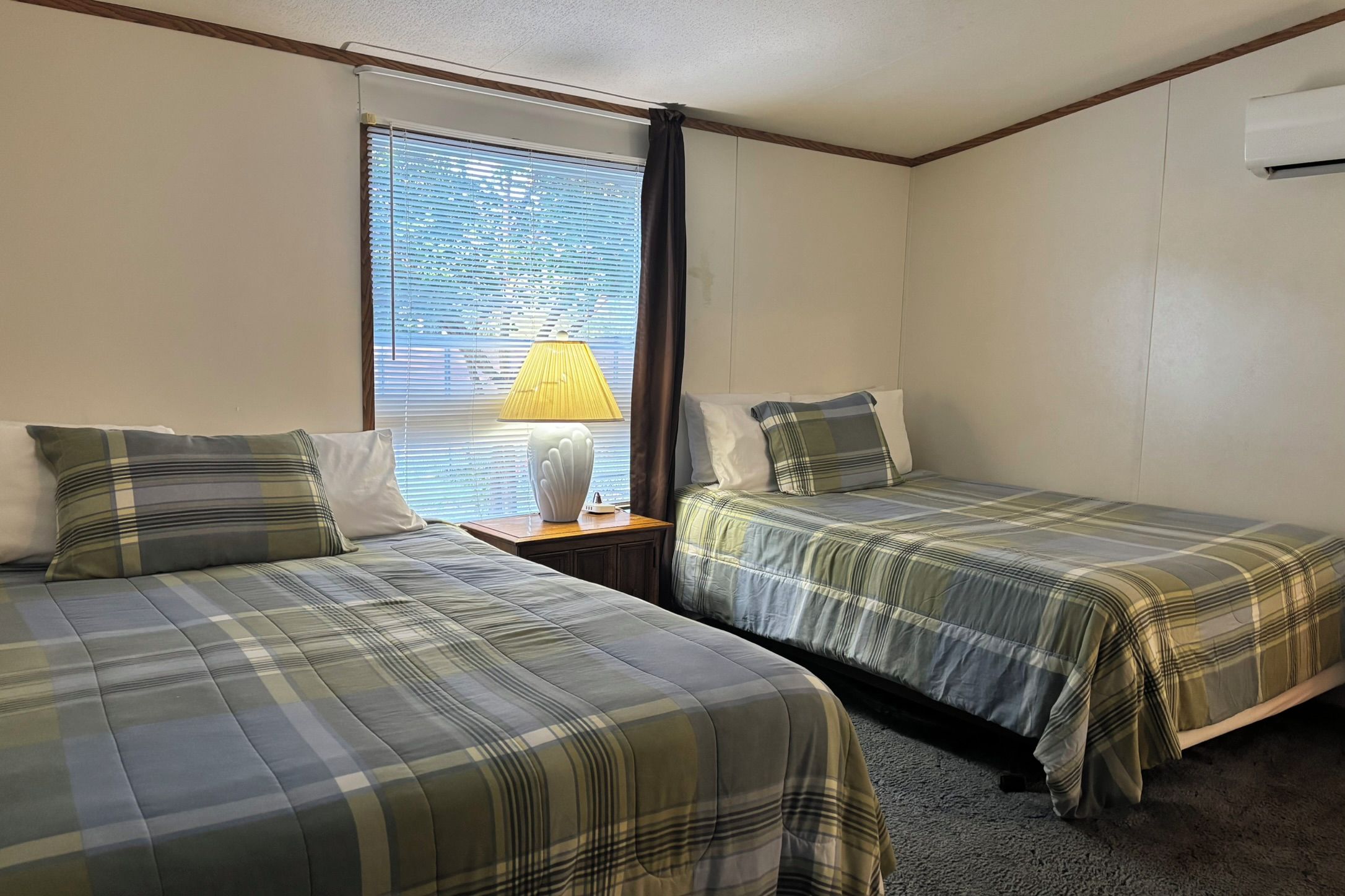 A room with two beds, a window with blinds, a lamp on a nightstand, and an air conditioning unit.