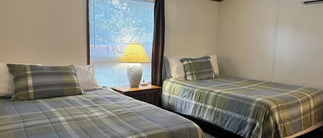Une chambre avec deux lits, une fenêtre avec des stores, une lampe sur une table de chevet et un appareil de climatisation.