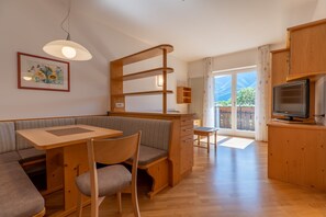 A cozy, well-lit room with a wooden dining table, cushioned seating, a television, and a view of the outdoors.