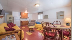 Een ruimtelijke woonkamer met een kitchenette, een eethoek met tafel en stoelen, een bank en een aan de muur bevestigde televisie.