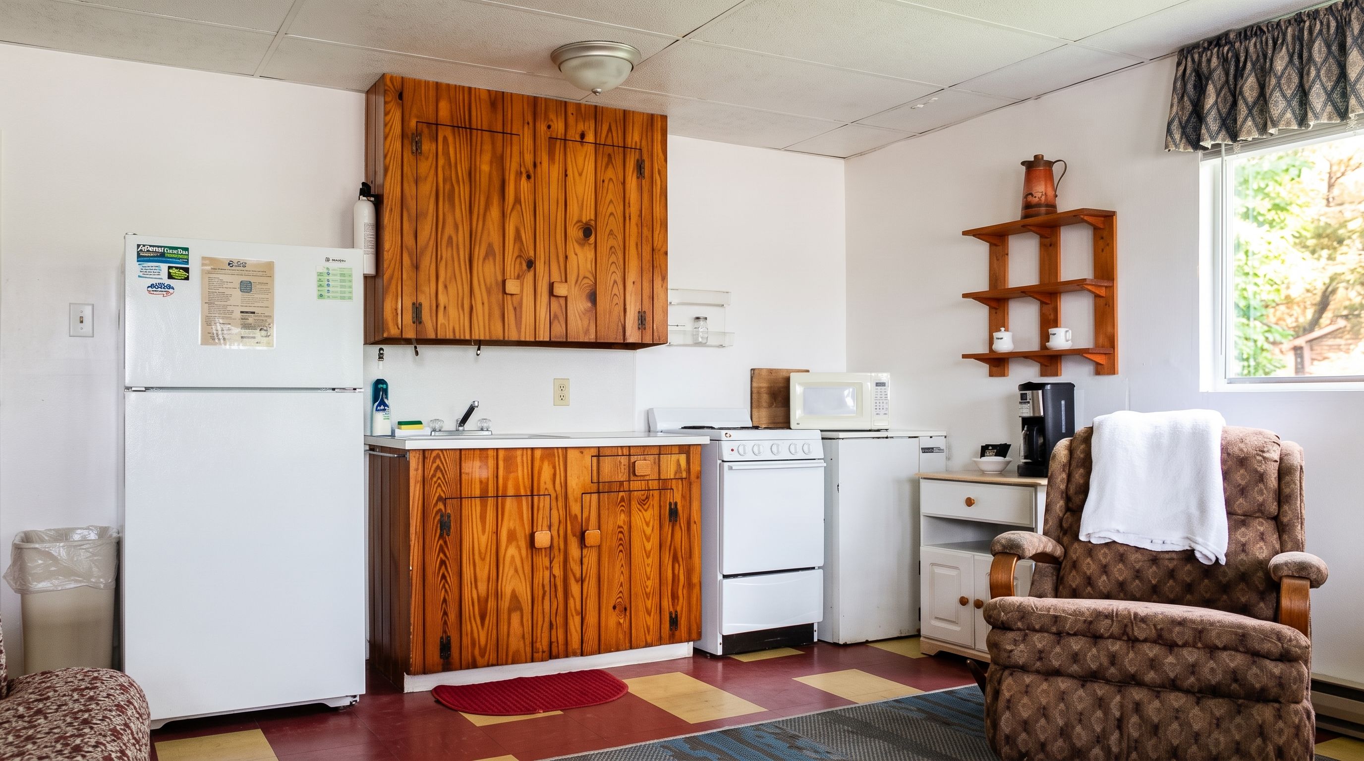 Une cuisine compacte avec des meubles en bois, un réfrigérateur blanc, une cuisinière, un four à micro-ondes et une machine à café. On y trouve une chaise sur laquelle est posée une serviette, ainsi qu’une fenêtre donnant sur un paysage verdoyant.