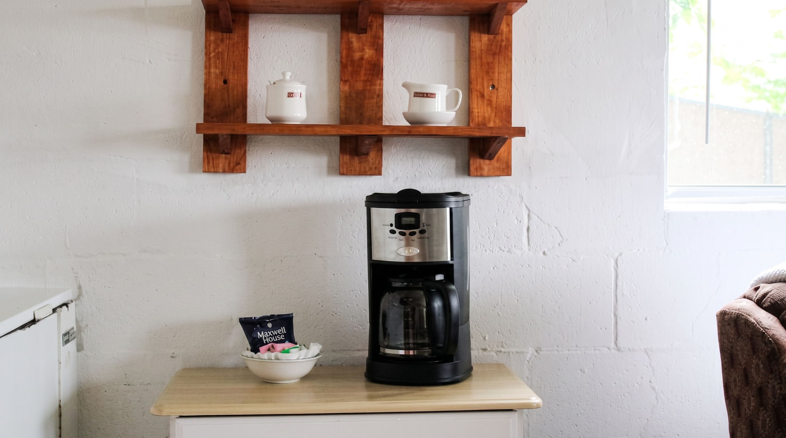 Een koffiezetapparaat op een houten tafel, met daaronder een wit kastje en erboven een houten plank met twee witte kopjes. Aan de rechterkant een raam.