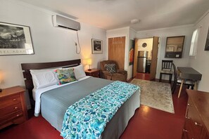 Un dormitorio con cama, mesitas de noche, una silla, un escritorio y una kitchenette.