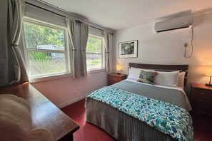 Ein Hotelzimmer mit Bett, Nachttischen, einem Fenster mit Vorhängen, einer Wandklimaanlage und einem gerahmten Bild.