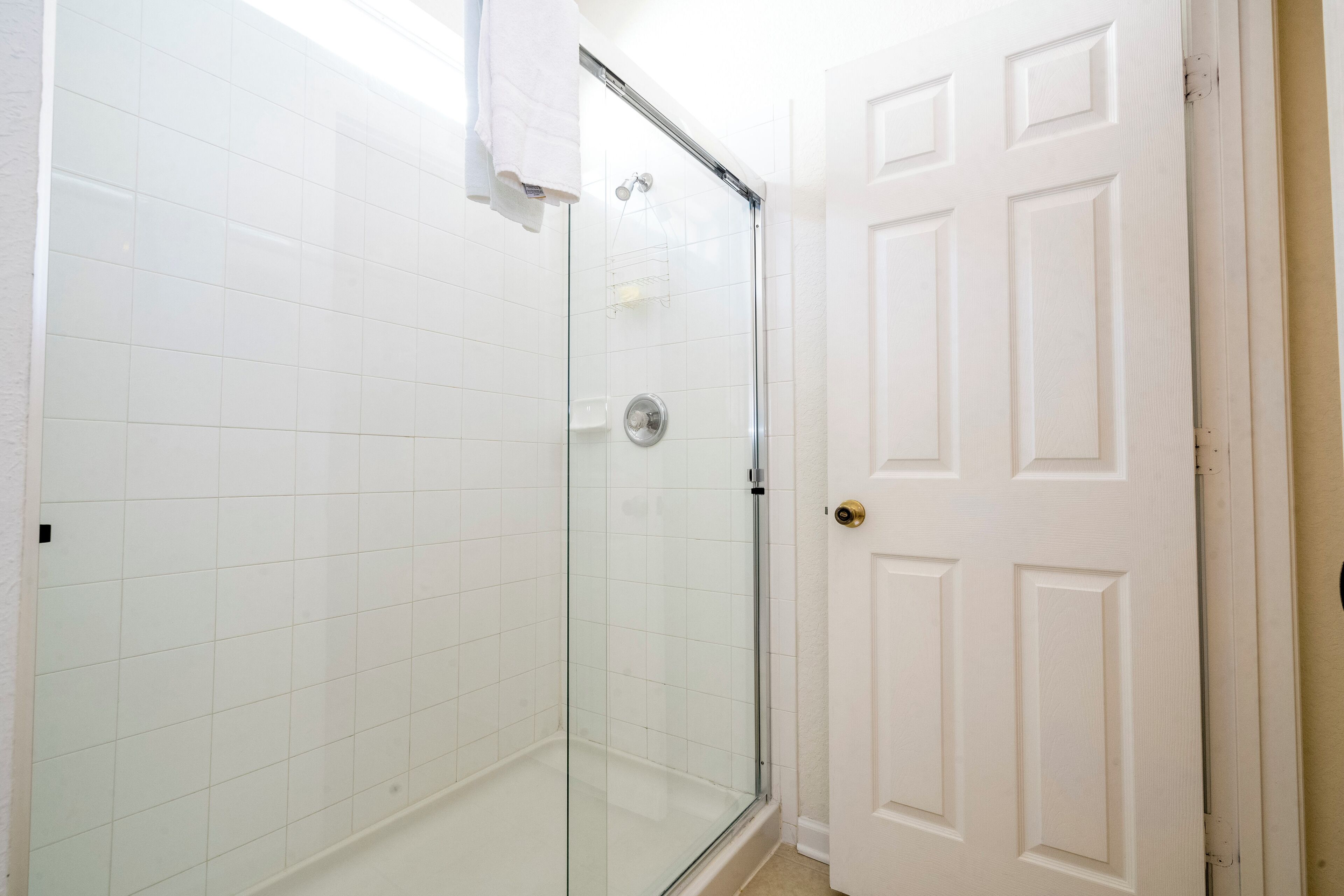 Un baño con paredes de azulejos blancos, cabina de ducha de vidrio y una puerta blanca.