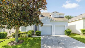 A residential house with a garage, a driveway, and a tree with autumn leaves. - Your Dream Family Vacation Home – 5BR, 5BA & Sun-Filled South-Facing Pool! (Kissimmee)