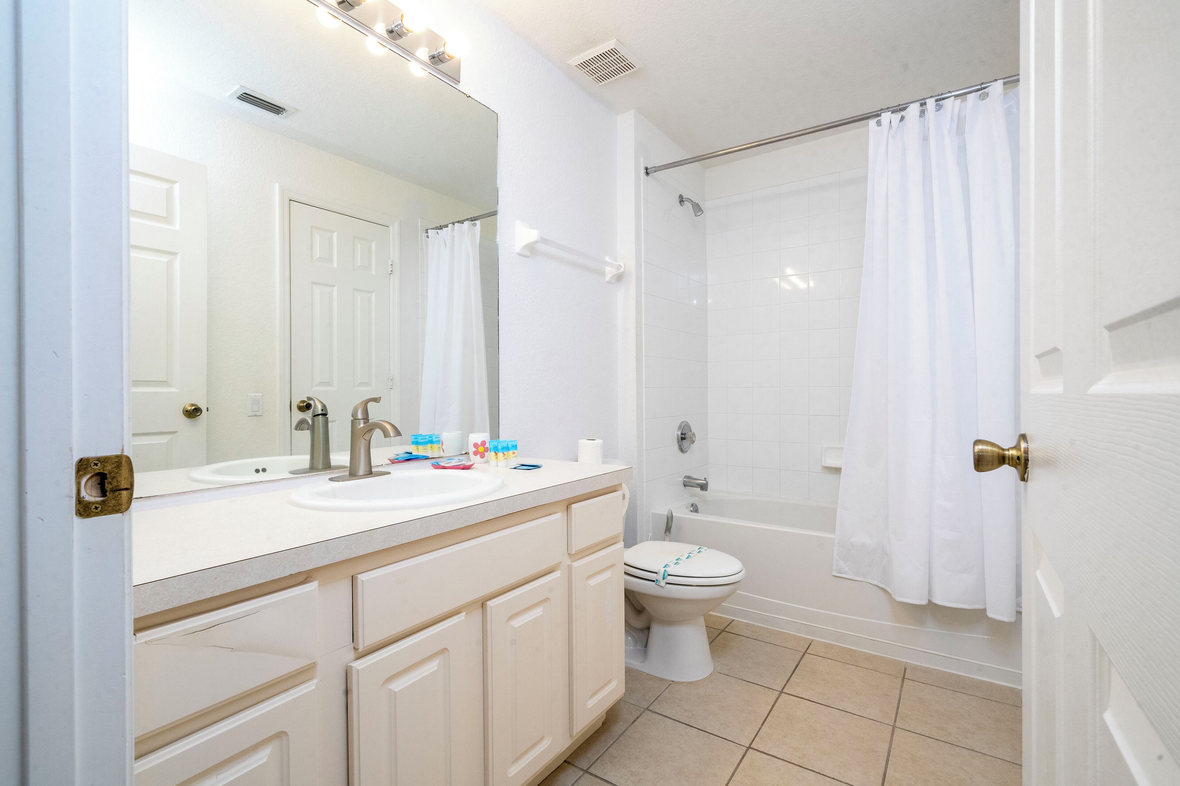 A bathroom with a white sink, mirror, and toilet, a shower with a curtain, and beige tiled flooring.