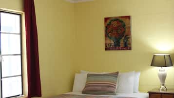 A room with a bed, a nightstand, a lamp, a window with curtains, and a floral painting on the wall.