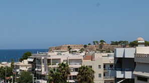 A coastal cityscape with a historic fort on a hill, modern buildings, and a clear view of the sea. - Domus Aries (Rethymno)