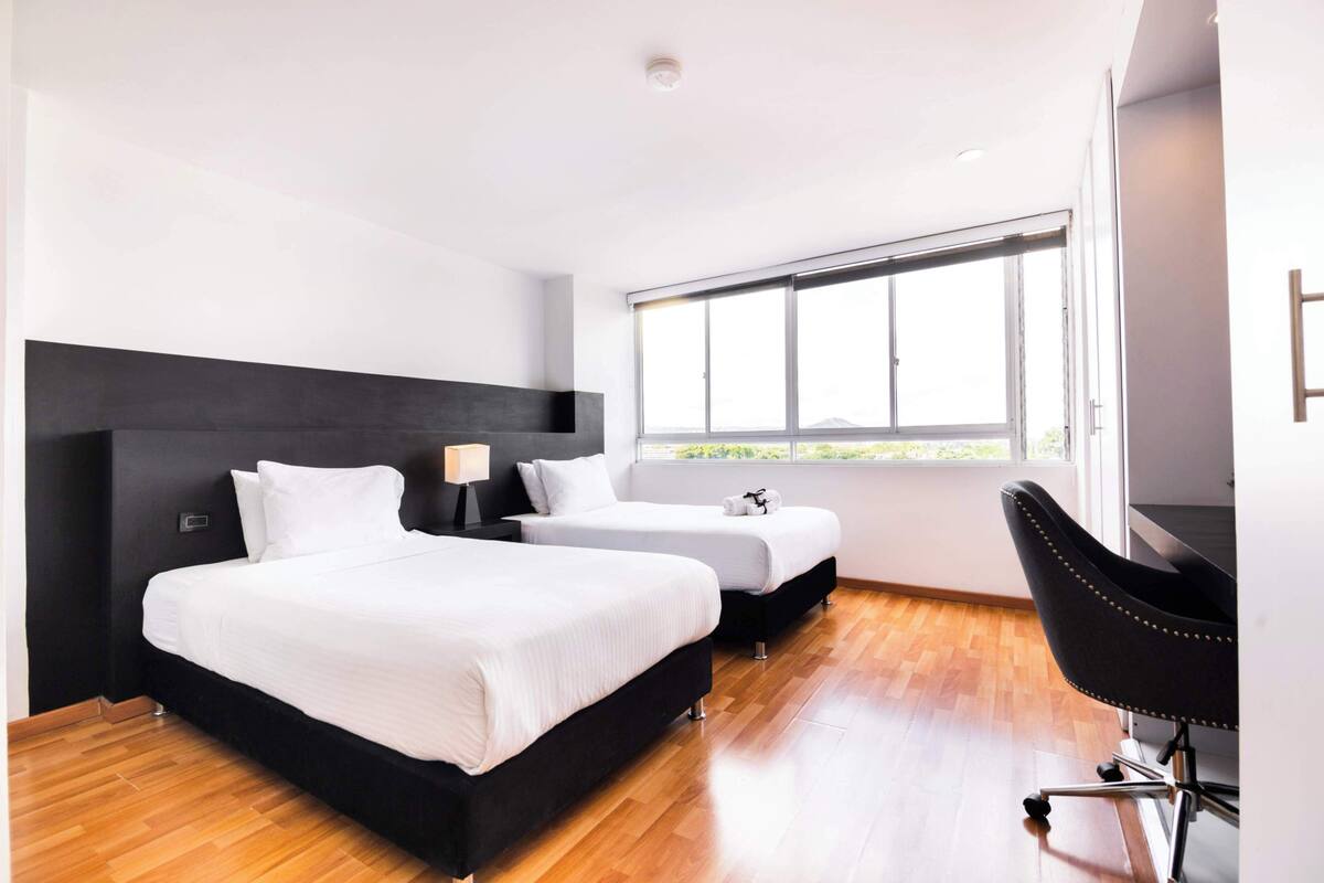 A modern hotel room with two beds, a black chair, and a large window providing a view of the outdoors.