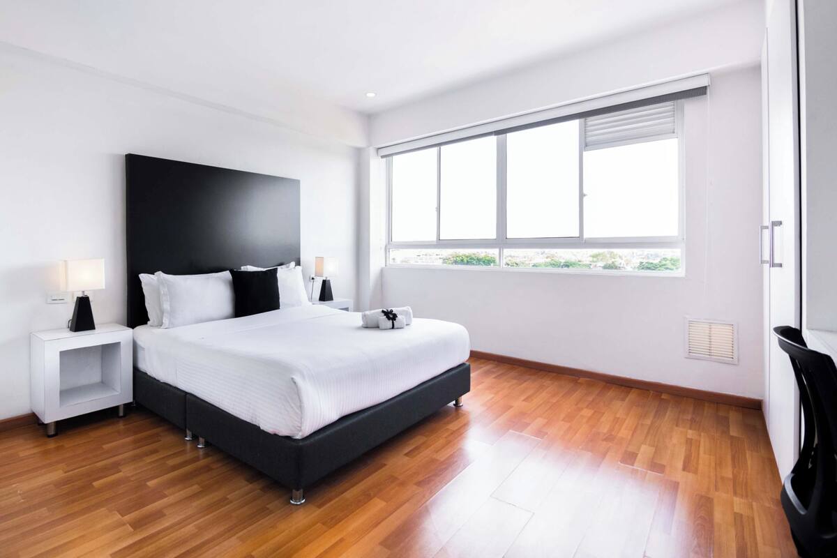 A modern bedroom with a large bed, white bedding, a black headboard, a nightstand with a lamp, and a large window with blinds.