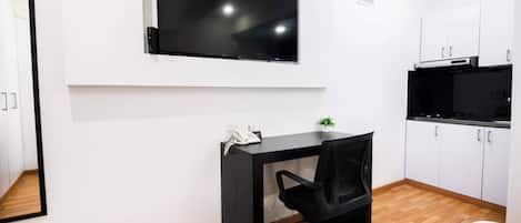 Una habitación moderna con un escritorio negro, una silla de oficina negra, un televisor de pantalla plana montado en la pared y armarios blancos.