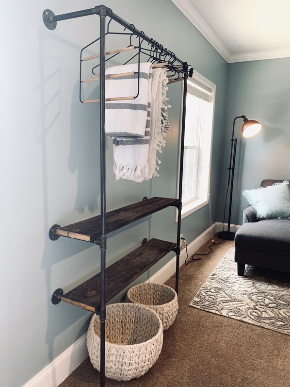 Ein Zimmer mit einer Metallregaleinrichtung an der Wand, einem Fenster mit Jalousien, einer Stehlampe, einem Bett mit einer hellen Tagesdecke und zwei gewebten Körben auf dem Boden.