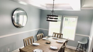 Un comedor con una mesa de madera dispuesta para cuatro personas, rodeada de sillas, una lámpara colgante y un espejo grande en la pared.