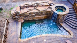 Un jacuzzi con una cascada de piedra, rodeado por una terraza de madera y un muro de piedra.