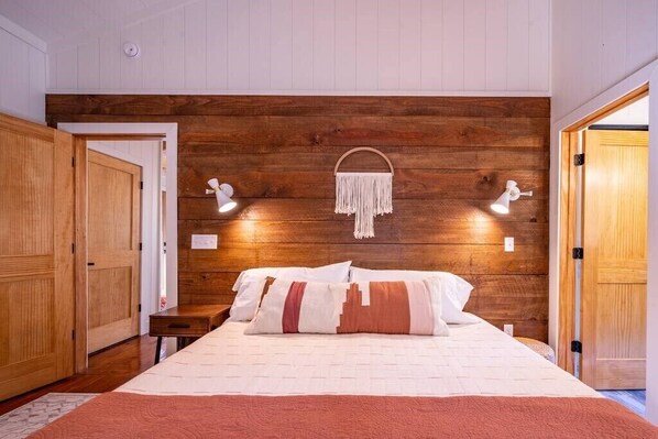 A bedroom with a wooden headboard, a bed with patterned bedding, bedside lamps, and a macrame wall hanging.