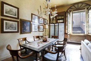 A dining room with a large table, chairs, a chandelier, framed pictures on the wall, and a view through an open window.