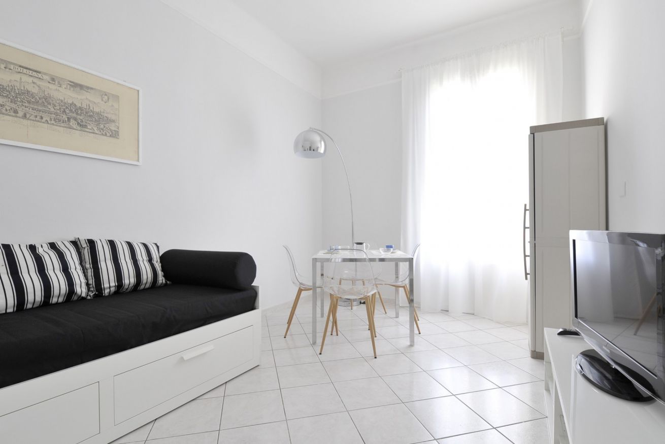 A modern, minimalist living room with a black and white striped sofa, a clear glass dining table with four chairs, a floor lamp, and a TV on a white cabinet.