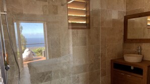 A bathroom with stone tiles, a wooden vanity, a glass shower enclosure, and a view of the ocean. - Charming chalet with large terrace and breathtaking sea view (Saint-Leu)