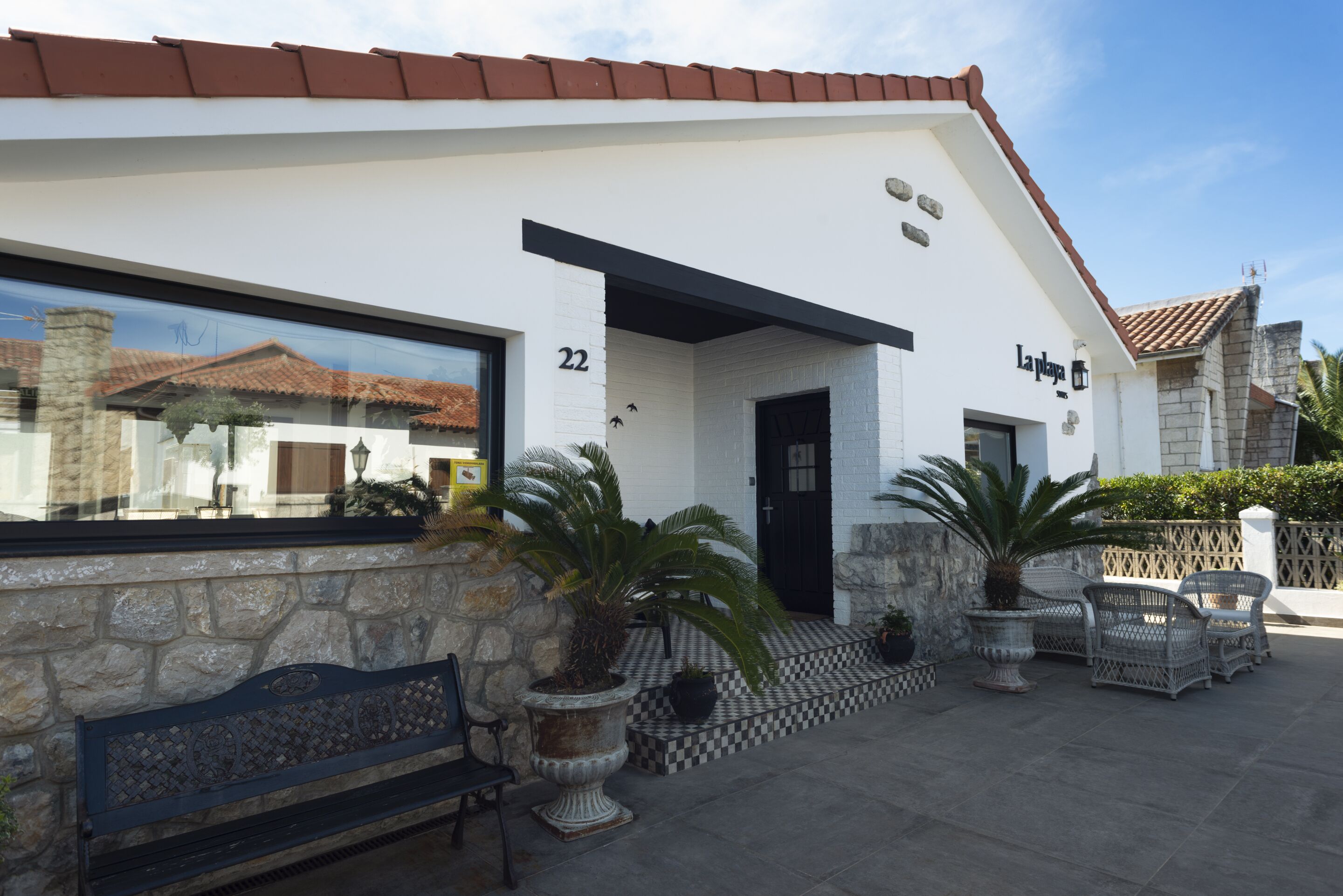 A building with a stone facade, numbered 22, and a black door. There are potted plants and wicker furniture outside.
