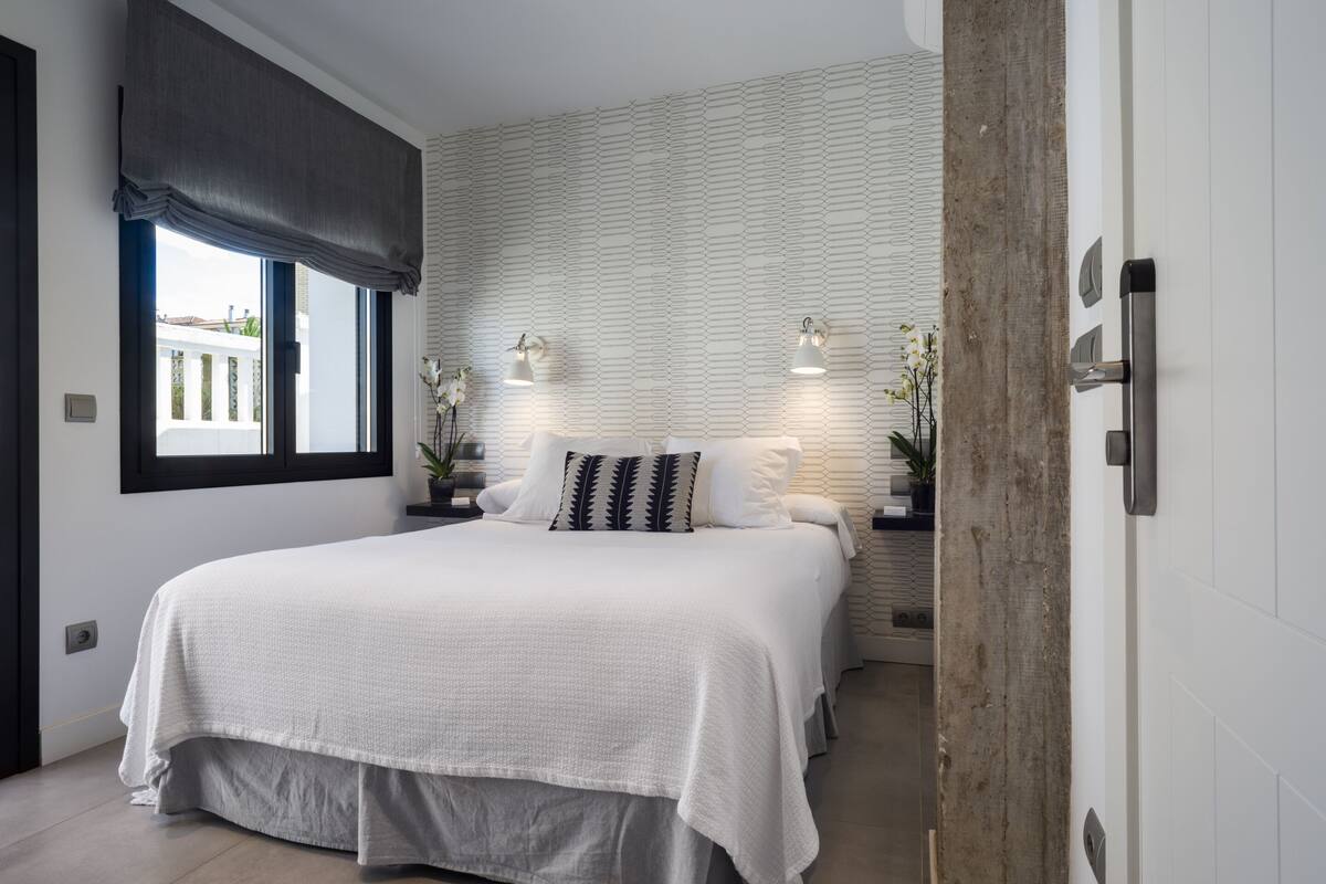 Un dormitorio moderno con una cama grande, dos mesitas de noche con lámparas, una ventana con cortina oscura y una pared con textura.