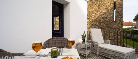 A dining area with a checkered floor, a wicker chair, a cushioned lounge chair, a table with a plate of food, two glasses of beer, and a candle.