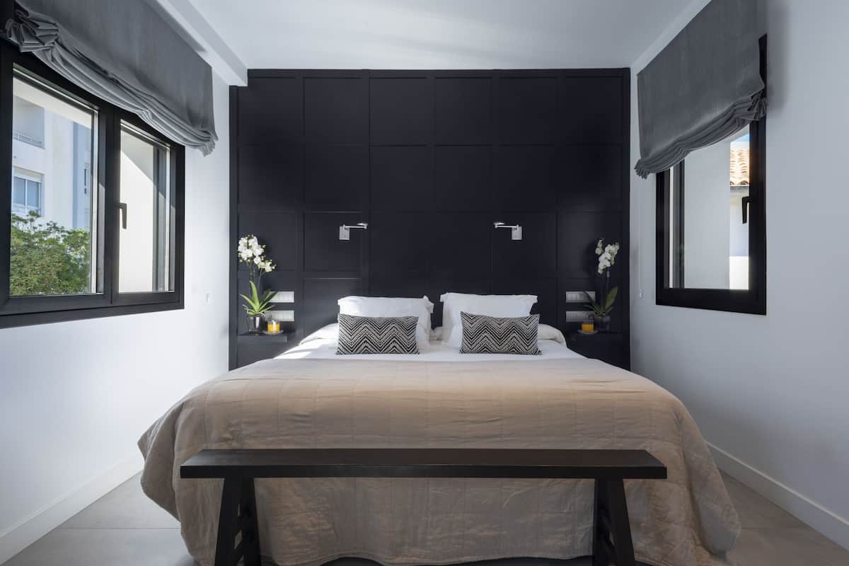 Un dormitorio moderno con una cama grande, cabecera oscura, mesitas de noche con lámparas y vistas al exterior.