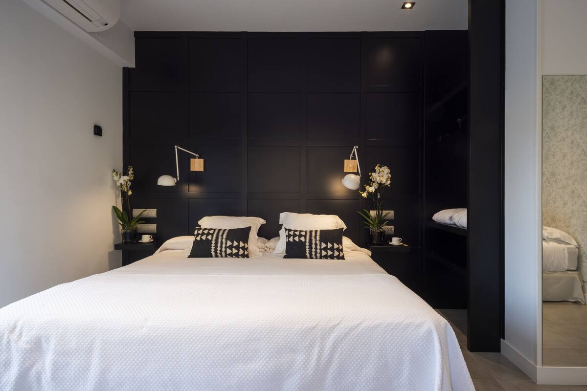 Un dormitorio moderno con una cama grande, dos mesitas de noche, un cabecero oscuro y una pequeña planta en maceta.