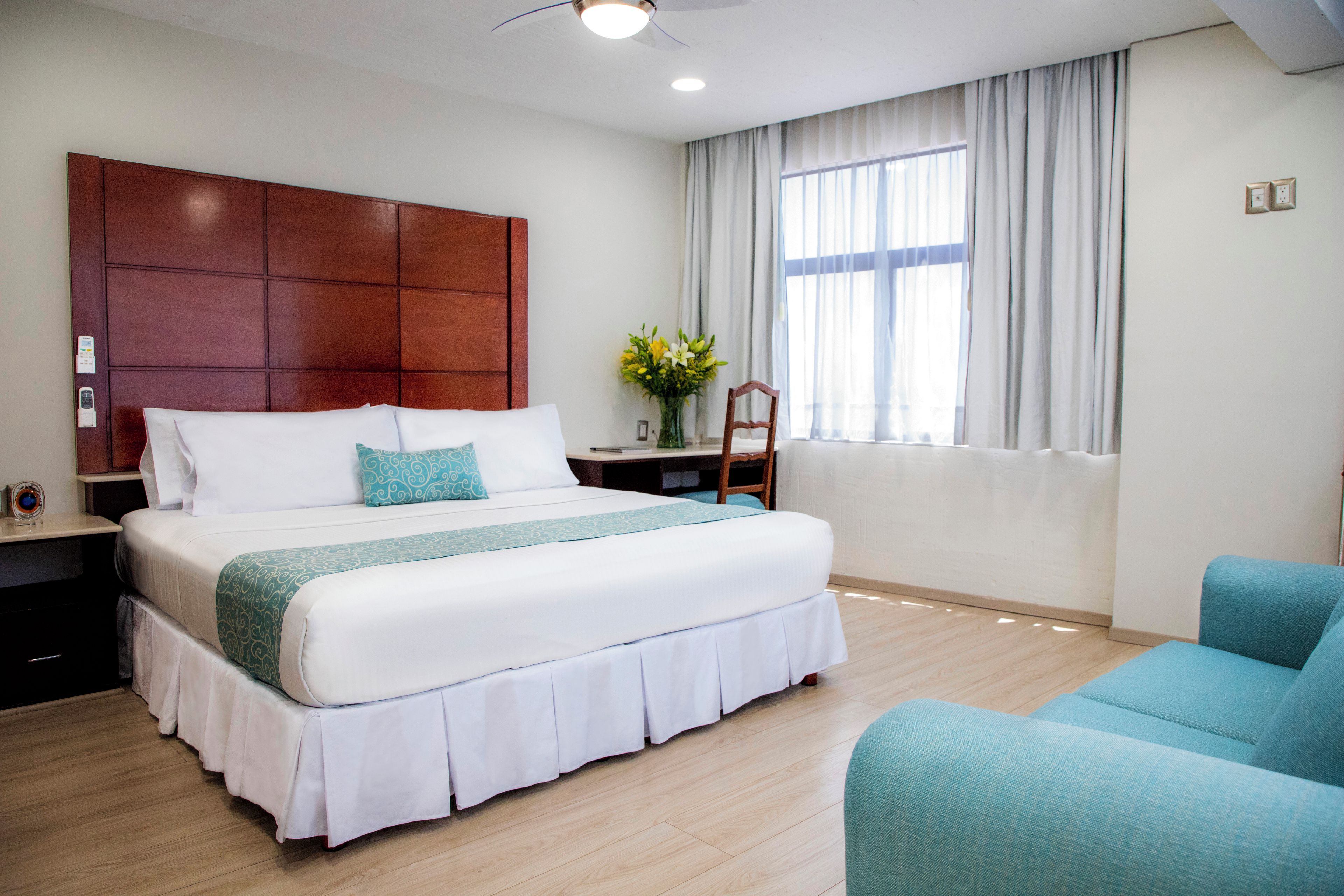 A hotel room with a large bed, a desk with a chair, a window with curtains, and a ceiling fan.