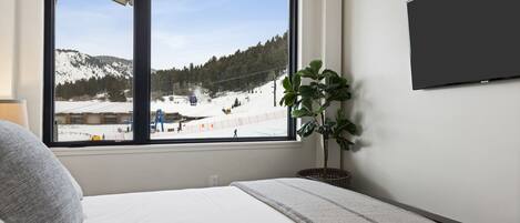 A bedroom with a large window overlooking a snowy landscape and a ski slope.