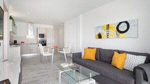 A modern living room with a glass coffee table, grey sofa with yellow cushions, and a dining area with white chairs. - Casa Mercedes, Apt 1 - Sea View and Terrace, Puerto del Carmen - Lanzarote (Puerto del Carmen)