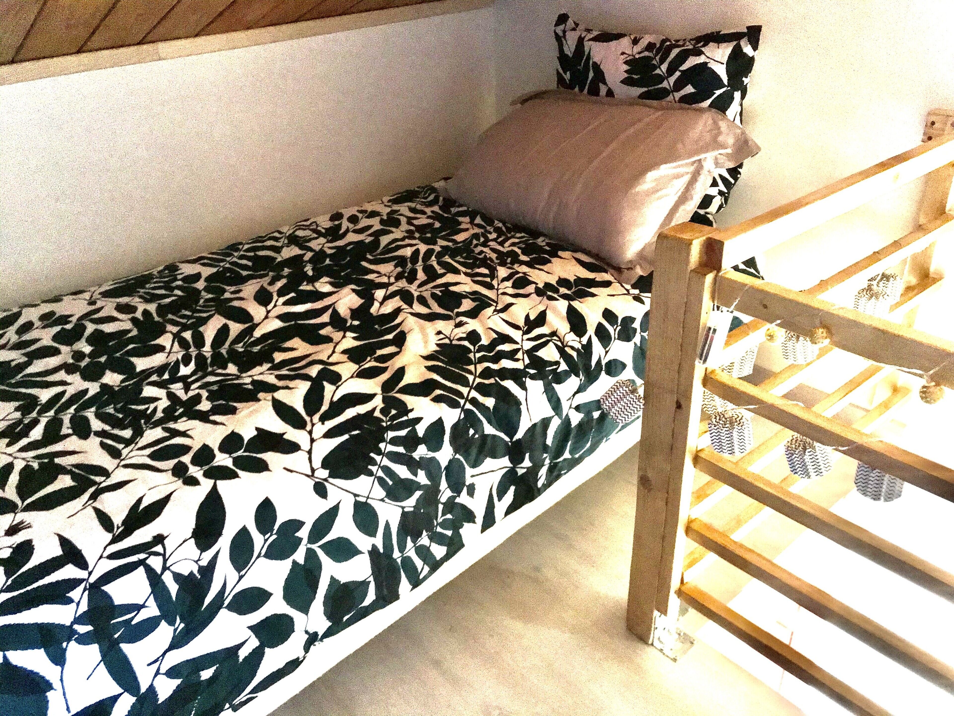 A single bed with a leaf-patterned bedspread, a wooden headboard, and a white wall.
