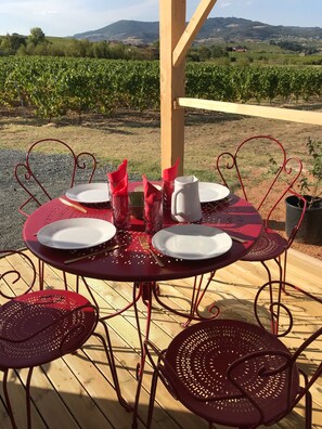 Une table dressée à l’extérieur, avec des chaises en métal rouge, une pergola en bois et un vignoble en arrière-plan.