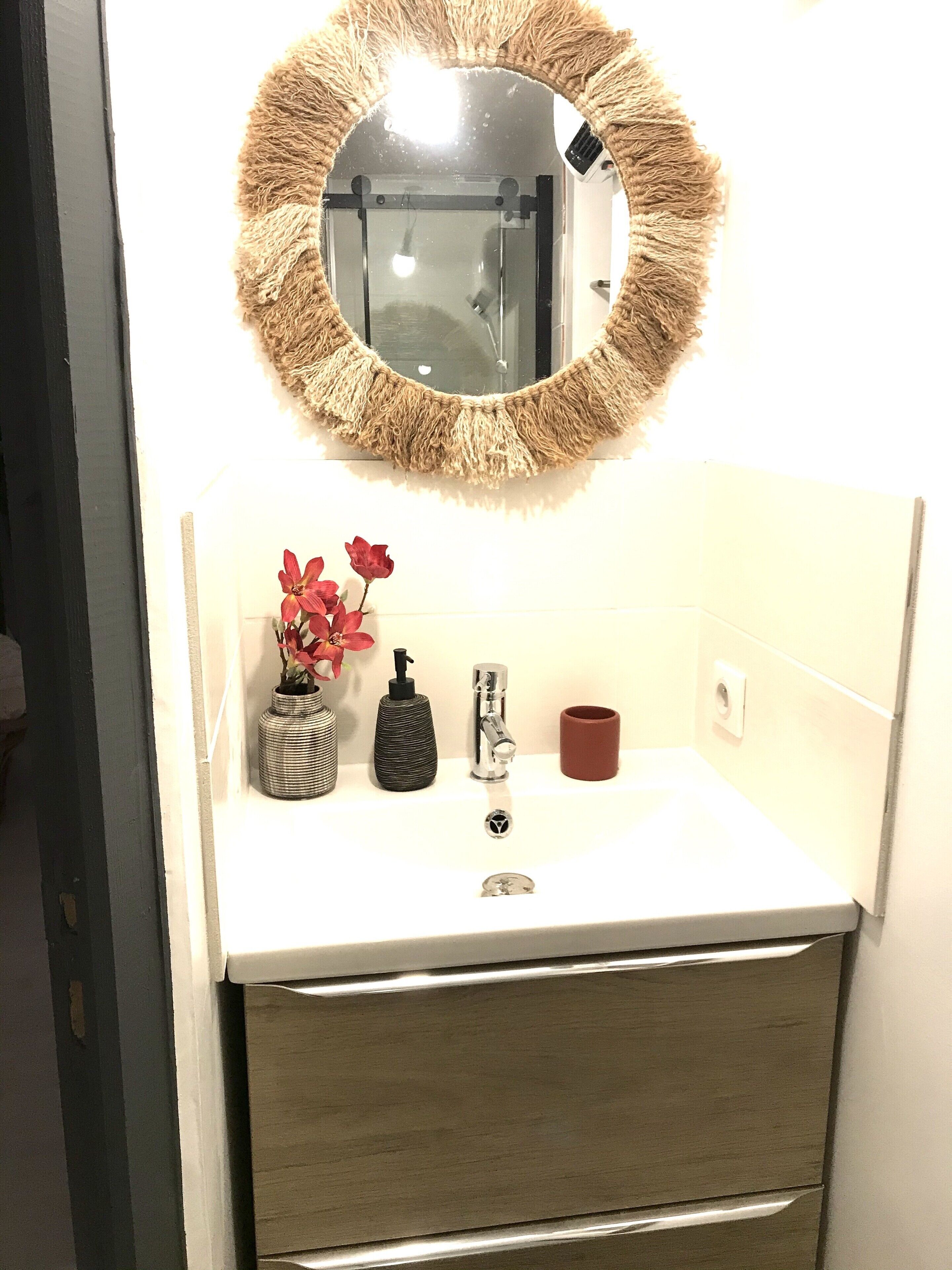 A bathroom with a white sink, a round mirror with a textured frame, a red vase with flowers, a black soap dispenser, and a brown mug.