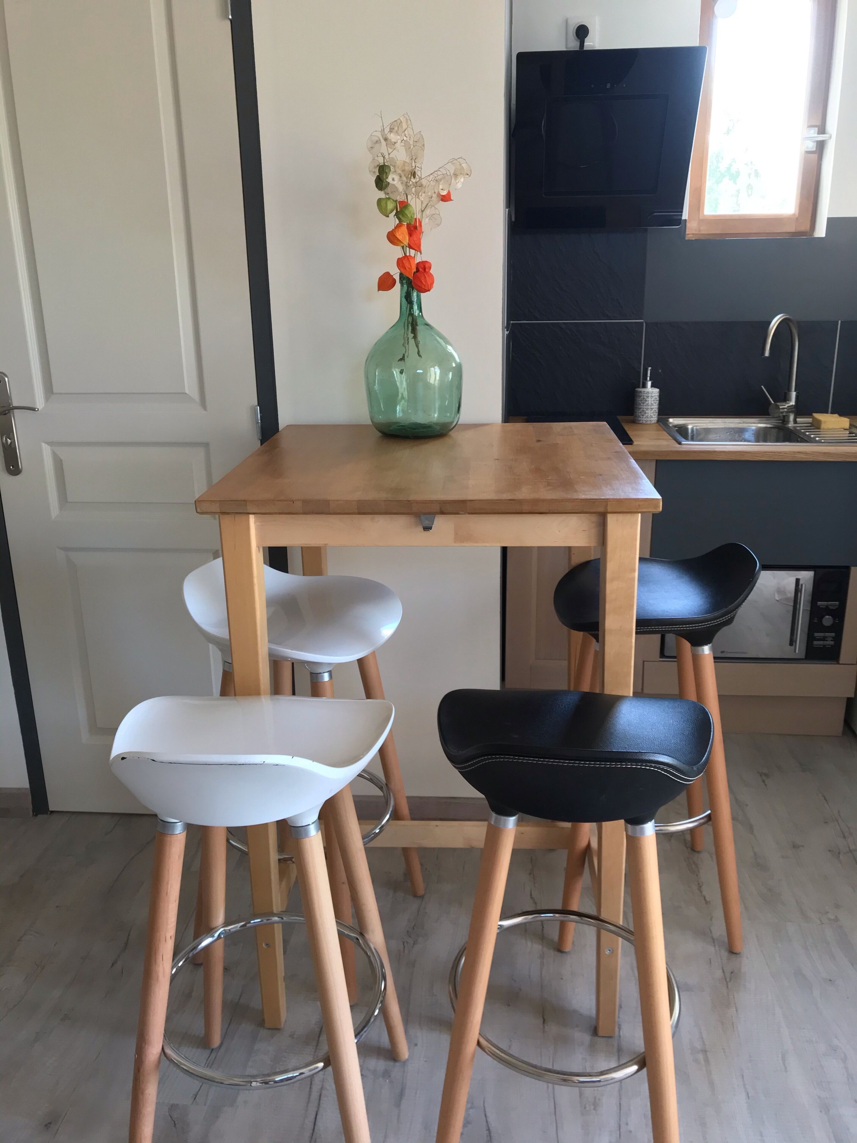 Una cocina con una mesa de madera, dos taburetes de bar, un florero con flores, un televisor negro y una ventana con vistas a árboles.