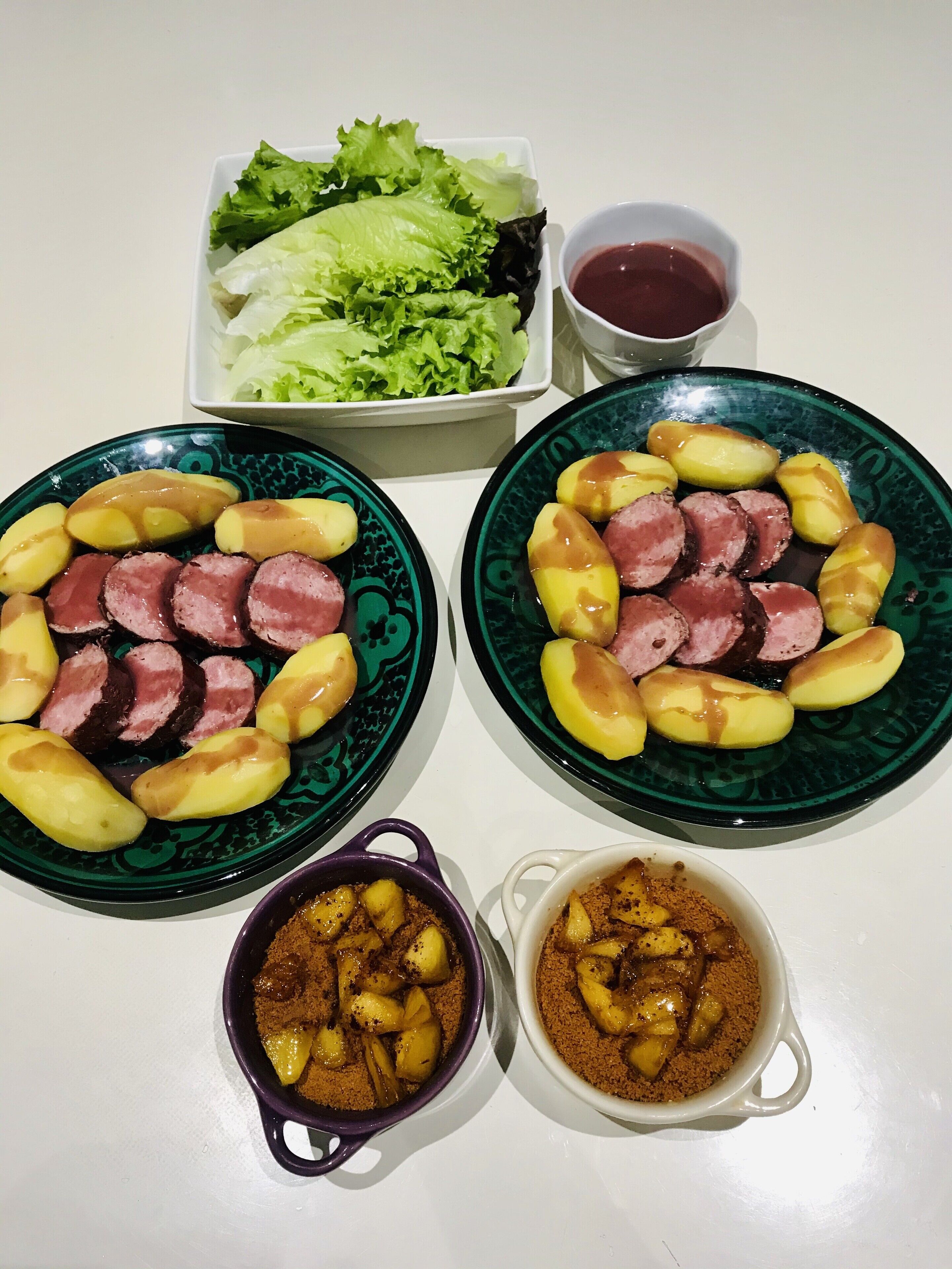 Deux assiettes de viande et de pommes de terre coupées en tranches, un saladier de salade et deux petits bols de sauce sont posés sur une table.