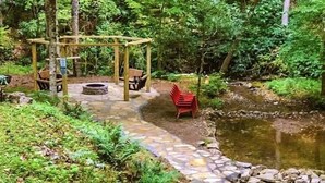 Un chemin de pierres mène à un belvédère en bois doté de bancs, le tout étant entouré d’une végétation luxuriante et d’un petit étang.