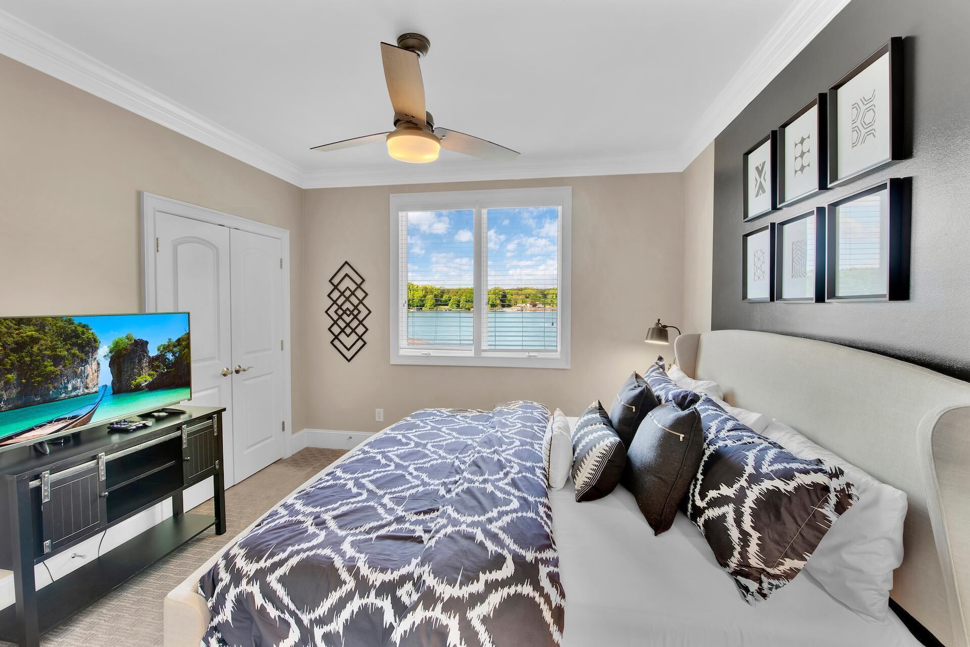 A bedroom with a bed, a TV, a ceiling fan, a window with a view, and framed pictures on the wall.
