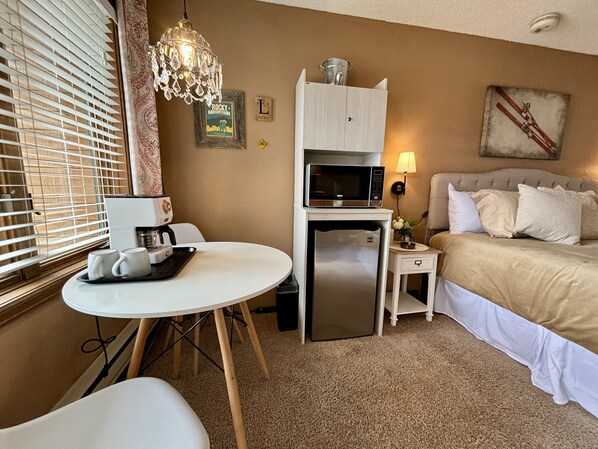 A hotel room with a bed, a small table with a coffee maker, a microwave, a refrigerator, a lamp, and a decorative wall piece.