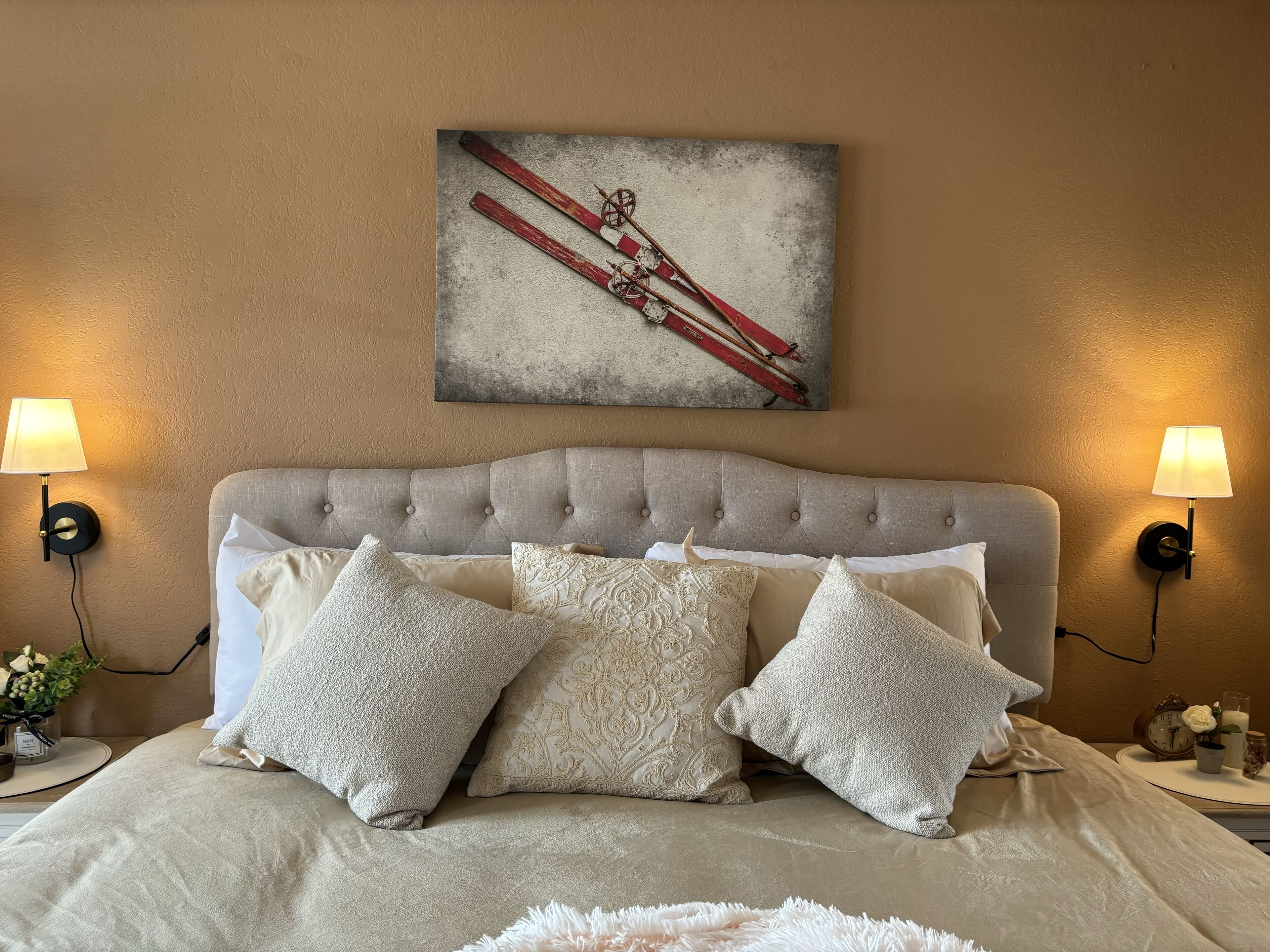 A bedroom with a tufted headboard, multiple pillows, two wall-mounted lamps, and a painting of skis on the wall.