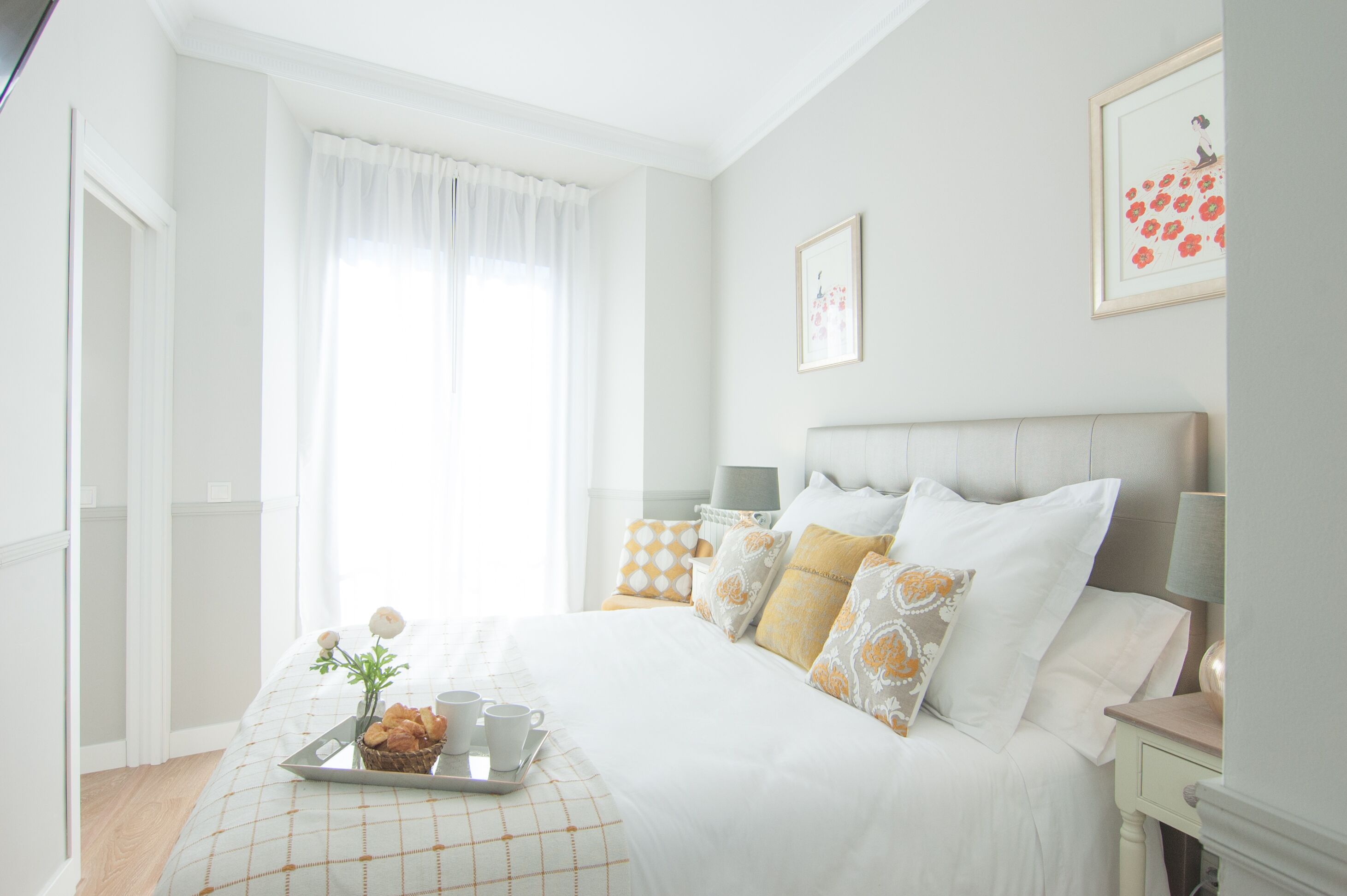 Un dormitorio ordenado con cama, mesitas de noche, una bandeja con tazas y una cesta, además de una ventana con cortinas.