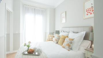 Un dormitorio ordenado con cama, mesitas de noche, una bandeja con tazas y una cesta, además de una ventana con cortinas.